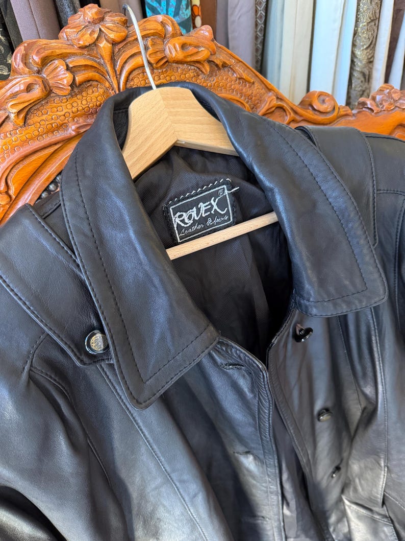 May include: A black leather jacket with a notched collar and button closures. The jacket is hanging on a wooden hanger. The label reads "ROVEX Leather Fashions".