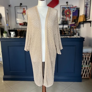 May include: A beige open-front cardigan with a loose, open-knit design. The long-sleeved sweater is displayed on a mannequin. The cardigan is a neutral colour and has a relaxed fit, suitable for layering. The background includes a shop interior.