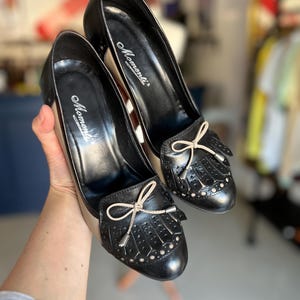 May include: A pair of black leather pumps with a beige interior. The shoes feature a bow and fringe detail on the toe, with small decorative perforations. The heels are approximately 2 inches high. The brand name "Momenti" is visible on the insole.