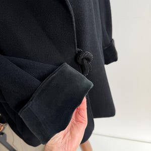 Pu&ograve; includere: Cappotto nero con tessuto strutturato e polsino in velluto a contrasto. Il cappotto presenta un'esclusiva chiusura annodata con dettaglio a nappa. La manica &egrave; ripiegata per rivelare il polsino. Il cappotto &egrave; esposto su un supporto di legno.