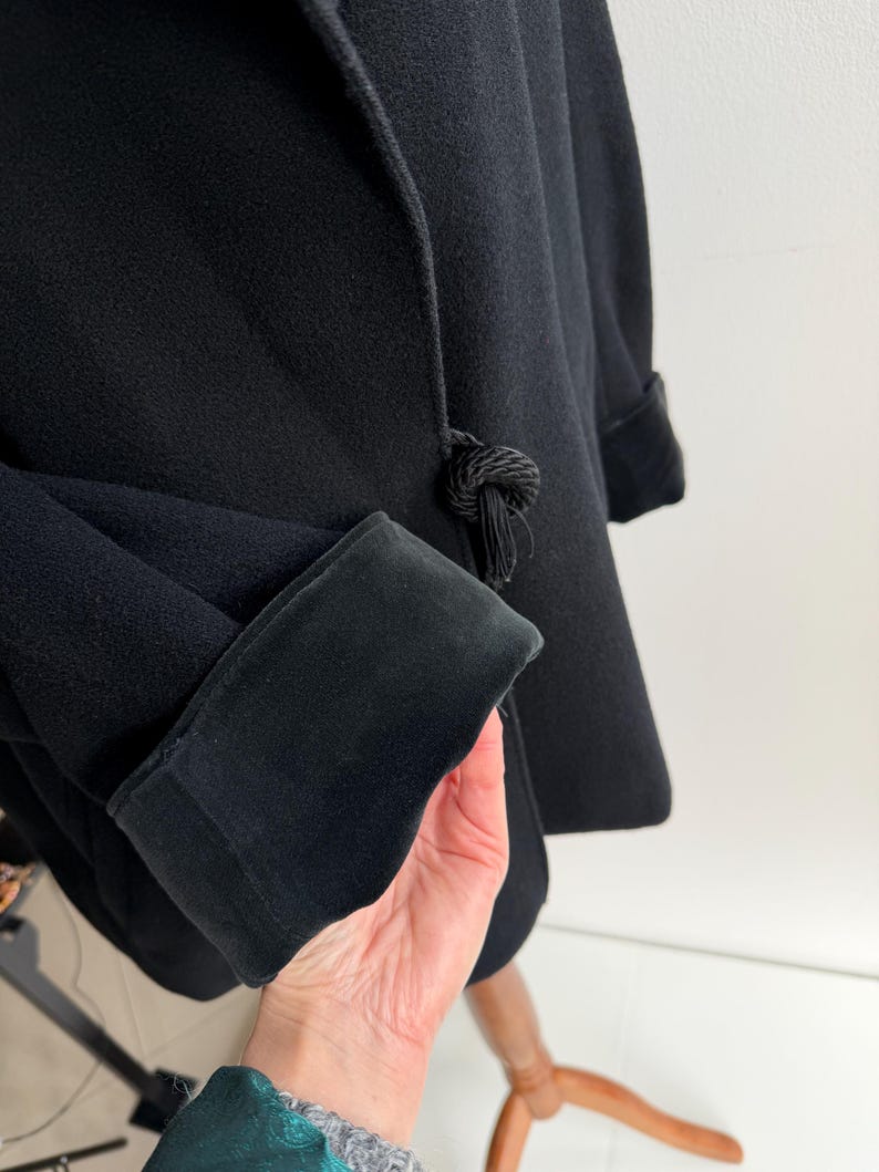 Pu&ograve; includere: Cappotto nero con tessuto strutturato e polsino in velluto a contrasto. Il cappotto presenta un'esclusiva chiusura annodata con dettaglio a nappa. La manica &egrave; ripiegata per rivelare il polsino. Il cappotto &egrave; esposto su un supporto di legno.