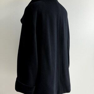 Pu&ograve; includere: Un cappotto nero in lana con ampio colletto e maniche lunghe. Il cappotto &egrave; esposto su una gruccia di legno su sfondo bianco. Le maniche hanno un dettaglio con polsino ripiegato.