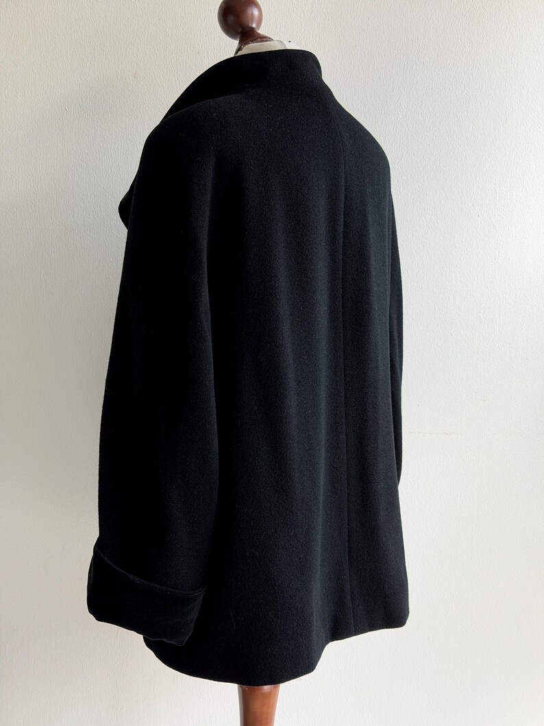 Pu&ograve; includere: Un cappotto nero in lana con ampio colletto e maniche lunghe. Il cappotto &egrave; esposto su una gruccia di legno su sfondo bianco. Le maniche hanno un dettaglio con polsino ripiegato.