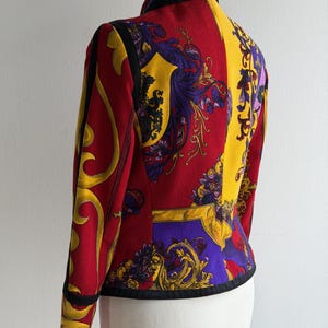 May include: A vibrant, patterned jacket in red, yellow, and purple. The jacket features a high collar and black trim. The design includes ornate, baroque-style motifs. The jacket is displayed on a mannequin, showcasing its tailored fit and intricate details.