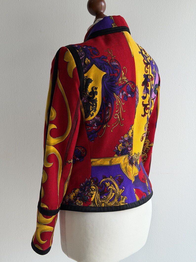 May include: A vibrant, patterned jacket in red, yellow, and purple. The jacket features a high collar and black trim. The design includes ornate, baroque-style motifs. The jacket is displayed on a mannequin, showcasing its tailored fit and intricate details.