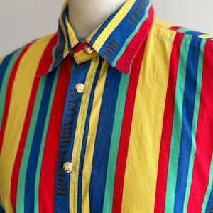 May include: A colorful button-up shirt with vertical stripes in red, yellow, blue, and green. The collar and cuffs feature the text "ROYALTY" in black. Gold-colored buttons secure the shirt.