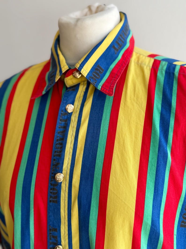 May include: A colorful button-up shirt with vertical stripes in red, yellow, blue, and green. The collar and cuffs feature the text "ROYALTY" in black. Gold-colored buttons secure the shirt.