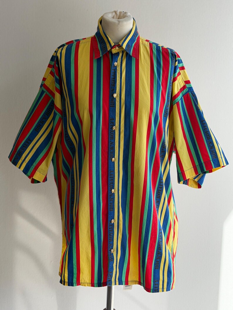 May include: A short-sleeved button-down shirt with vertical stripes in red, yellow, blue, and green. The shirt has a classic collar and gold-colored buttons. The fabric appears to be cotton or a similar material.
