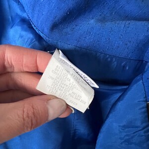 May include: A close-up of a blue fabric label with white text. The text reads: "Esterno Exterieur Outside Sen 69K Viscosa 4X Poliammide Naylon Viskose Polyamide Nylon Peseta Soie Polyakid Poliamide Styl Setos Seda".