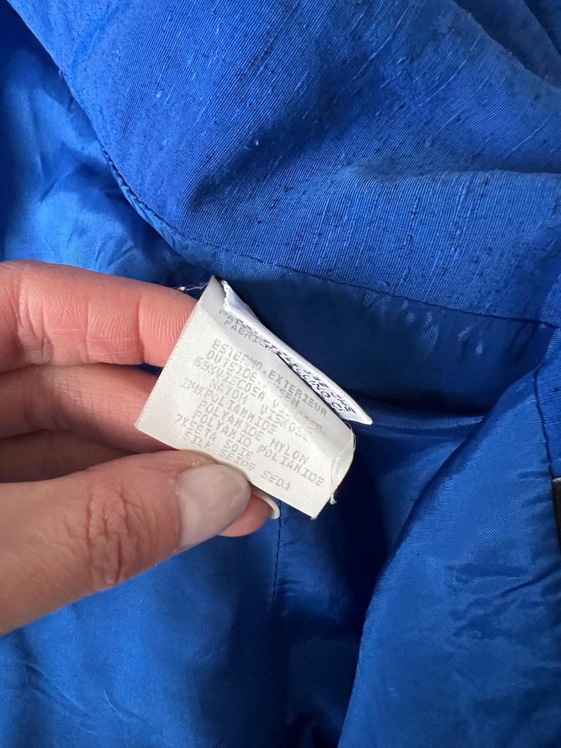 May include: A close-up of a blue fabric label with white text. The text reads: "Esterno Exterieur Outside Sen 69K Viscosa 4X Poliammide Naylon Viskose Polyamide Nylon Peseta Soie Polyakid Poliamide Styl Setos Seda".