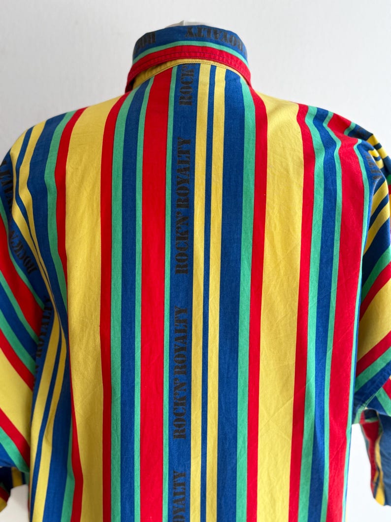 May include: A colorful striped shirt with vertical stripes in red, yellow, blue, and green. The text "ROCK'N ROYALTY" is printed vertically on the back. The shirt has a button-down collar and short sleeves.