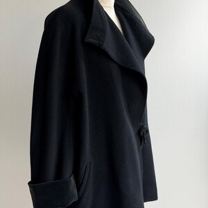 Pu&ograve; includere: Un cappotto nero in lana con un ampio colletto e un design drappeggiato unico. Il cappotto presenta un polsino ripiegato sulla manica e un dettaglio decorativo sul lato. Il cappotto &egrave; esposto su un manichino di legno.