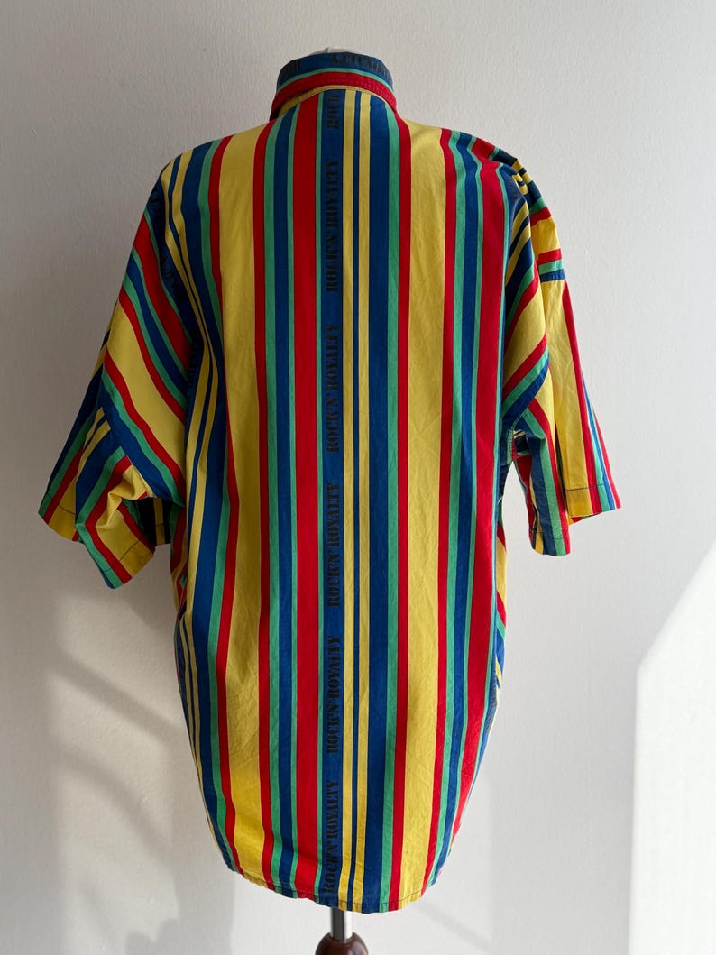 May include: A short-sleeved shirt with vertical stripes in yellow, red, blue, and green. The shirt has a button-down collar and the text "ROCK'S ROYALTY" printed vertically on the back.