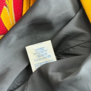 May include: Close-up of a black lining of a garment, with a tag visible. The tag has text in multiple languages, including "WOOLWOLLE" and care instructions. The garment's exterior features a colorful striped pattern in yellow, red, and pink.