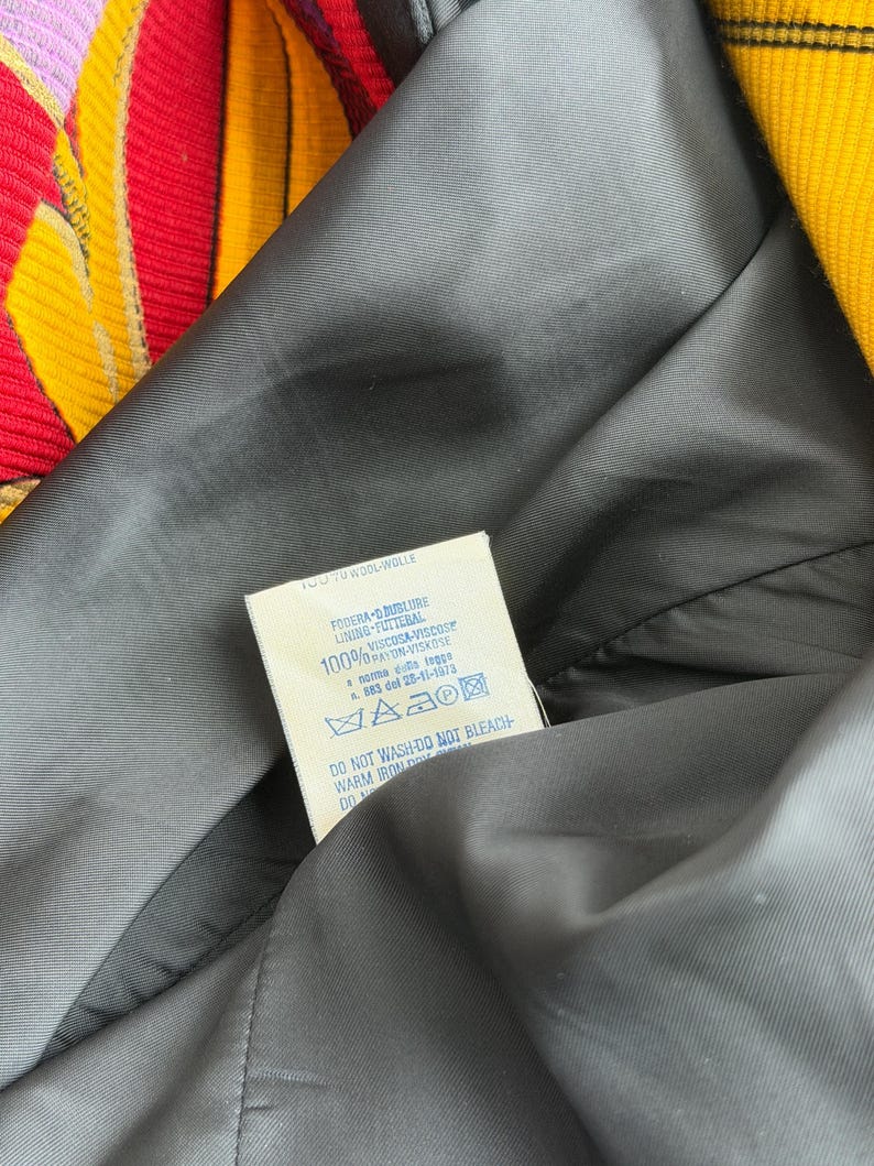 May include: Close-up of a black lining of a garment, with a tag visible. The tag has text in multiple languages, including "WOOLWOLLE" and care instructions. The garment's exterior features a colorful striped pattern in yellow, red, and pink.