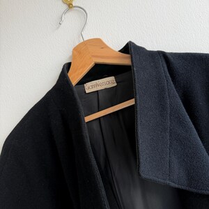 Pu&ograve; includere: Un cappotto nero di lana con un ampio colletto, appeso a una gruccia di legno. L'interno del cappotto &egrave; nero e un'etichetta con la scritta "Gianni Versace" &egrave; visibile. Il cappotto &egrave; appeso a un gancio di ottone su un muro bianco.