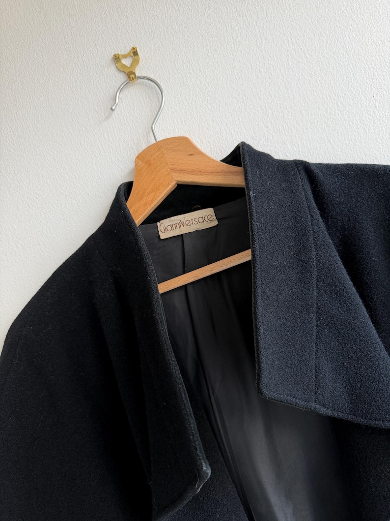 Pu&ograve; includere: Un cappotto nero di lana con un ampio colletto, appeso a una gruccia di legno. L'interno del cappotto &egrave; nero e un'etichetta con la scritta "Gianni Versace" &egrave; visibile. Il cappotto &egrave; appeso a un gancio di ottone su un muro bianco.