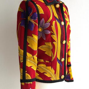 May include: A red jacket with a floral pattern featuring yellow leaves and purple flowers. The jacket has black trim, gold buttons, and a collared neckline. The jacket is displayed on a mannequin.