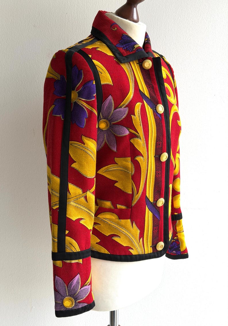 May include: A red jacket with a floral pattern featuring yellow leaves and purple flowers. The jacket has black trim, gold buttons, and a collared neckline. The jacket is displayed on a mannequin.