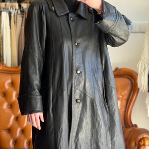 May include: A black leather coat with a button-up front, a collar, and long sleeves. The coat has a classic design and appears to be vintage. The coat is knee-length and has a slightly flared shape.