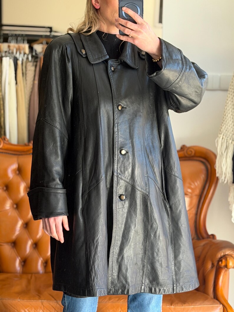 May include: A black leather coat with a button-up front, a collar, and long sleeves. The coat has a classic design and appears to be vintage. The coat is knee-length and has a slightly flared shape.