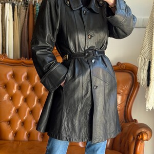 May include: Black leather trench coat with a belted waist, button closures, and a classic collar. The coat features a knee-length design and is worn with blue jeans. The coat has a vintage style.