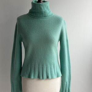 May include: A pale green turtleneck jumper. The jumper features long sleeves, a ribbed hem and cuffs, and a ribbed turtleneck. It is displayed on a mannequin.
