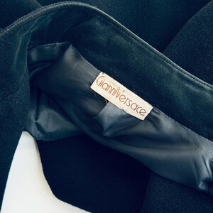 Pu&ograve; includere: Primo piano di un capo nero, possibilmente un cappotto o una giacca, con fodera scura. L'etichetta all'interno recita "Gianni Versace" in un carattere serif. Il tessuto sembra essere lana pesante o simile.