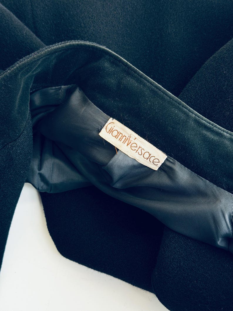 Pu&ograve; includere: Primo piano di un capo nero, possibilmente un cappotto o una giacca, con fodera scura. L'etichetta all'interno recita "Gianni Versace" in un carattere serif. Il tessuto sembra essere lana pesante o simile.