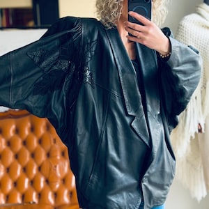 Könnte beinhalten: Schwarze Lederjacke mit großem floralem Design auf der Schulter. Die Jacke hat einen breiten Kragen und eine lockere Passform. Das Model trägt eine blaue Jeans. Die Jacke scheint Vintage zu sein.