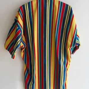 May include: A short-sleeved shirt with vertical stripes in yellow, red, blue, and green. The shirt has a button-down collar and the words "ROCK'S ROYALTY" are printed vertically on some of the stripes. The shirt is hanging on a wooden hanger.