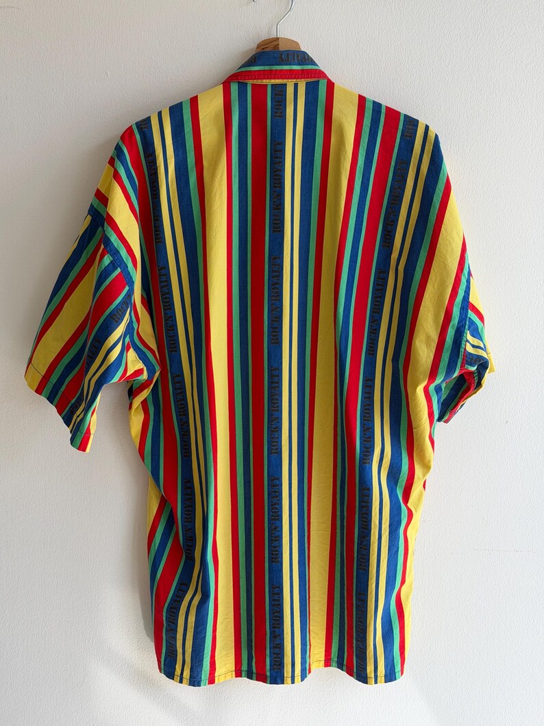 May include: A short-sleeved shirt with vertical stripes in yellow, red, blue, and green. The shirt has a button-down collar and the words "ROCK'S ROYALTY" are printed vertically on some of the stripes. The shirt is hanging on a wooden hanger.