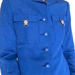 May include: A royal blue blazer with a button-up front and silver hardware. The blazer has two flap pockets with silver buckles and a silver button on each pocket flap. There are six silver buttons down the front of the blazer.
