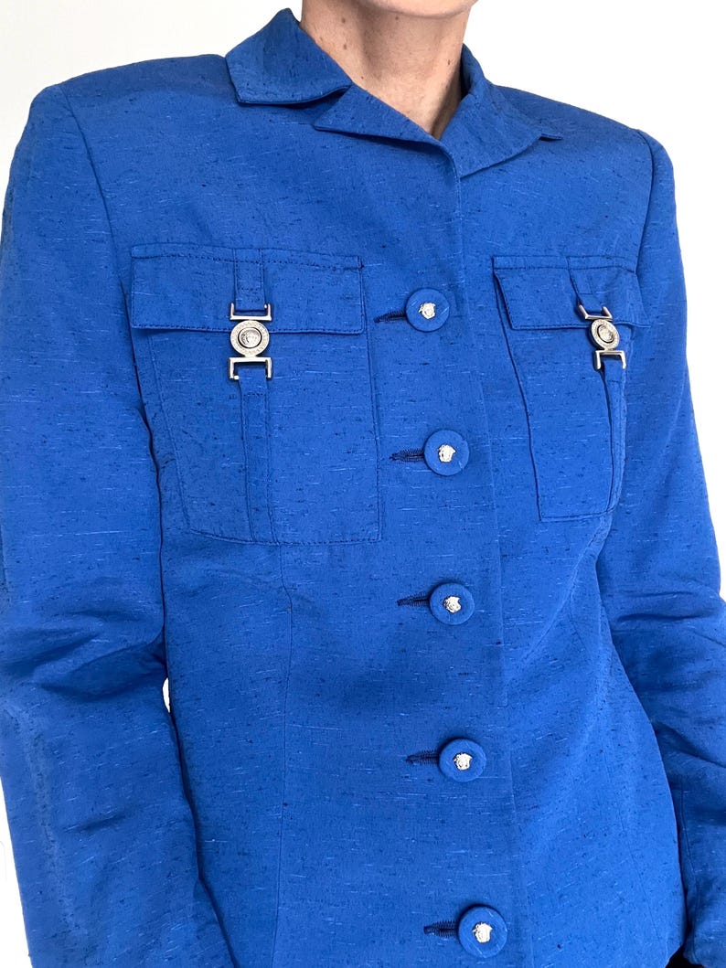 May include: A royal blue blazer with a button-up front and silver hardware. The blazer has two flap pockets with silver buckles and a silver button on each pocket flap. There are six silver buttons down the front of the blazer.