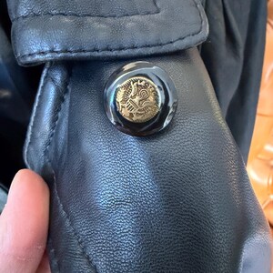 May include: Close-up of a black leather jacket detail. The jacket features a large, round button with a gold emblem. The button is set against the textured leather, and the stitching is visible.