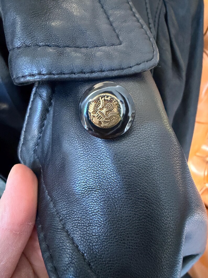 May include: Close-up of a black leather jacket detail. The jacket features a large, round button with a gold emblem. The button is set against the textured leather, and the stitching is visible.