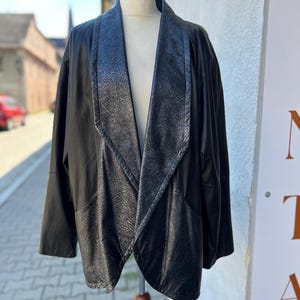 Peut inclure: Veste noire avec revers et panneaux avant texturés, présentée sur un mannequin. La veste présente un design unique avec une texture contrastée sur le revers et le devant, créant un look élégant. La veste est de couleur foncée.