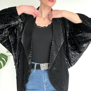 May include: Black sequined jacket with a velvet collar and lapel, paired with a black top and blue jeans. The jacket features a unique design with a decorative belt.
