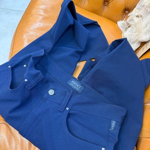May include: A pair of solid navy blue trousers featuring a button closure, a waistband label reading Versace Jeans Couture, and small silver-toned metal rivets on the front pockets. A small rectangular tag with the word Versace is on the pocket.
