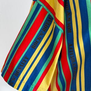 May include: A short-sleeved shirt with vertical stripes in red, yellow, blue, and green. The text "ROCK'N ROYALTY" is printed vertically in black on the sleeve. The shirt has a classic, casual style.