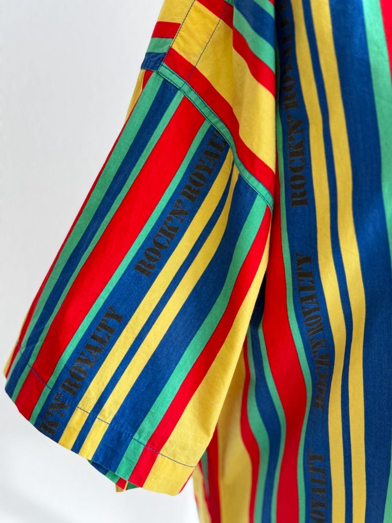 May include: A short-sleeved shirt with vertical stripes in red, yellow, blue, and green. The text "ROCK'N ROYALTY" is printed vertically in black on the sleeve. The shirt has a classic, casual style.
