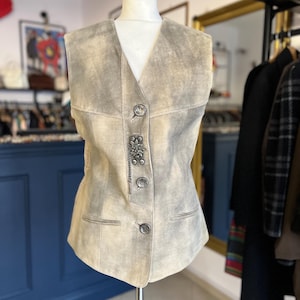 Op de afbeelding: Een beige suède vest met een V-hals en decoratieve metalen knopen. Het vest heeft een getailleerde pasvorm, met twee kleine zakken en een gedetailleerde metalen sluiting. Het kledingstuk wordt op een mannequin gepresenteerd.
