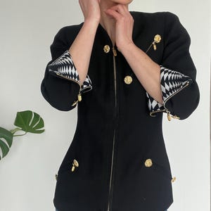 May include: Black jacket with gold-tone buttons and zipper. The jacket features a geometric black and white pattern on the cuffs. The jacket has a tailored fit and is a classic style. The jacket is a versatile piece that can be dressed up or down.