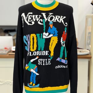 May include: A black long-sleeve sweater featuring a crew neck with yellow and green trim. The front displays colorful graphic text reading NEW YORK, SNOW, FLORIDE, STYLE, and adidas, alongside illustrations of people skiing in bright clothing.
