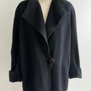 Pu&ograve; includere: Un cappotto nero con ampio colletto e polsini. Il cappotto ha un'esclusiva chiusura a nodo e una silhouette su misura. Il tessuto sembra essere una morbida misto lana. Il cappotto &egrave; esposto su un manichino.