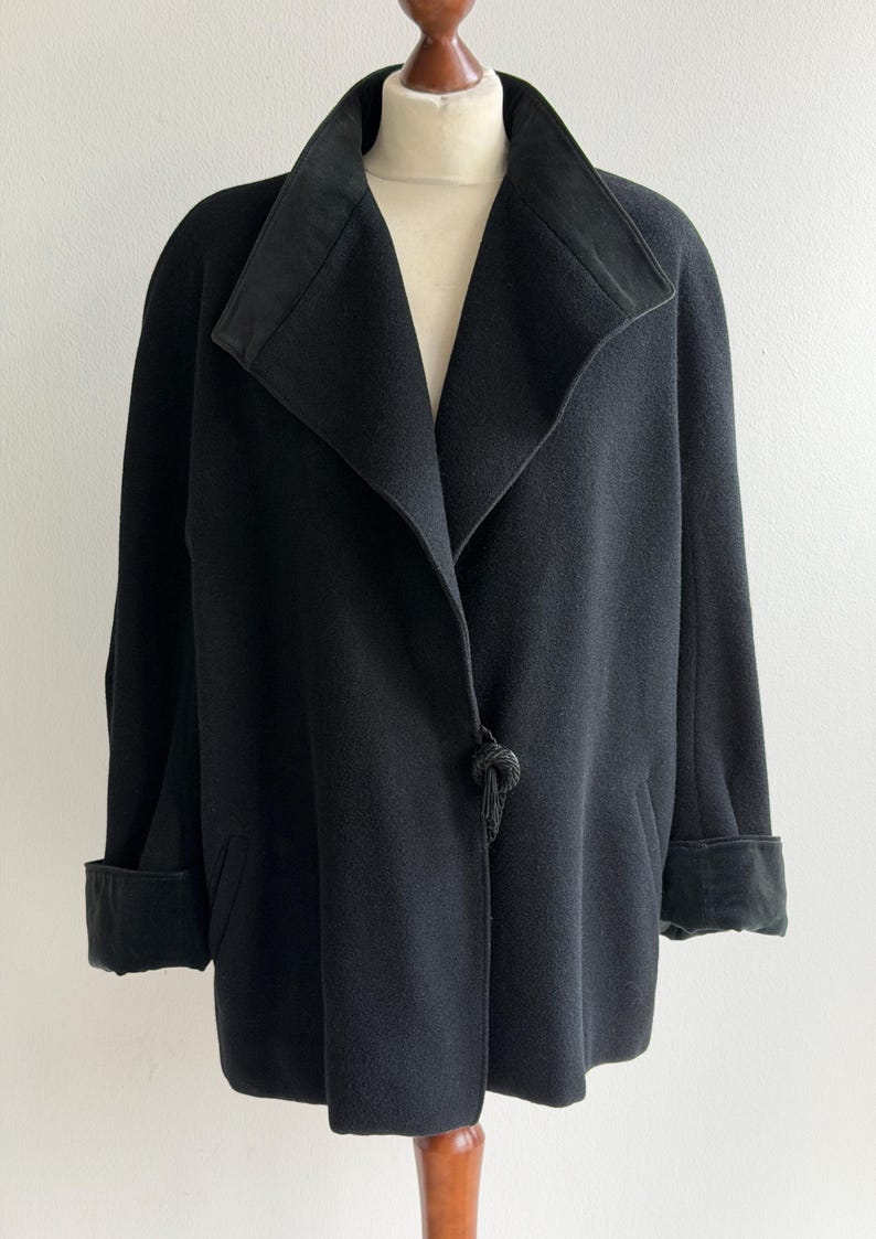 Pu&ograve; includere: Un cappotto nero con ampio colletto e polsini. Il cappotto ha un'esclusiva chiusura a nodo e una silhouette su misura. Il tessuto sembra essere una morbida misto lana. Il cappotto &egrave; esposto su un manichino.