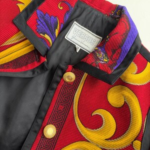 May include: A close-up of a vintage Versus Gianni Versace jacket. The jacket features a red, black, yellow, and purple patterned design with gold buttons. The label "VERSUS Gianni Versace" is visible.