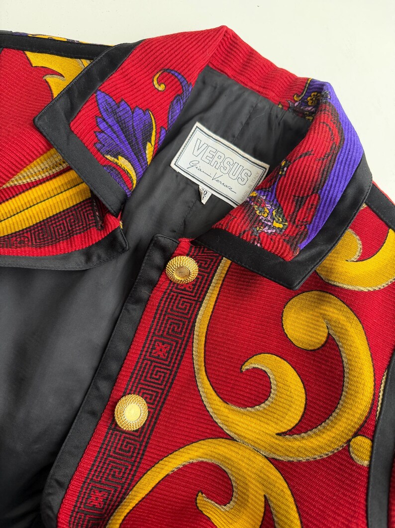 May include: A close-up of a vintage Versus Gianni Versace jacket. The jacket features a red, black, yellow, and purple patterned design with gold buttons. The label "VERSUS Gianni Versace" is visible.