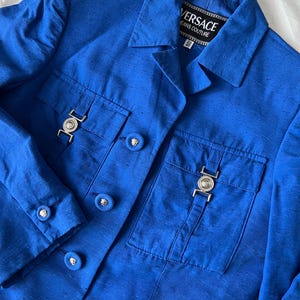 May include: A blue blazer with a button closure and two flap pockets. The blazer has a silver metal buckle on each pocket and the label "VERSACE JEANS COUTURE" on the collar.