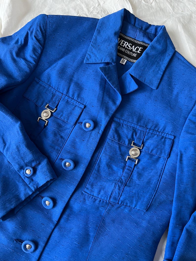 May include: A blue blazer with a button closure and two flap pockets. The blazer has a silver metal buckle on each pocket and the label "VERSACE JEANS COUTURE" on the collar.